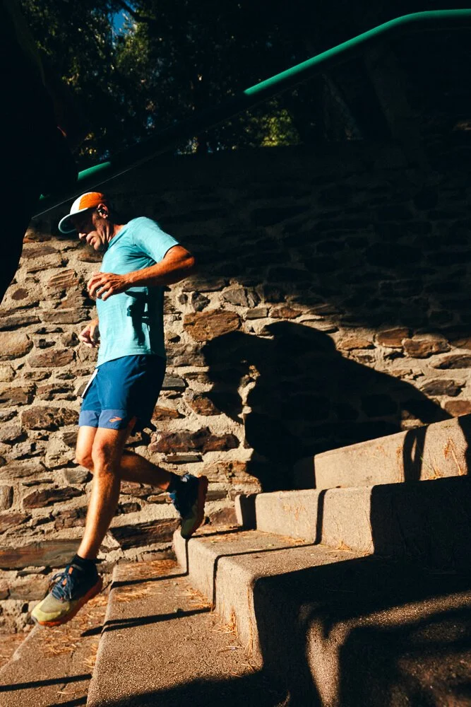 A man wearing a light blue shirt, navy shorts, and a white and orange cap is walking down concrete stairs outdoors during daytime, casting a shadow on a stone wall behind him.