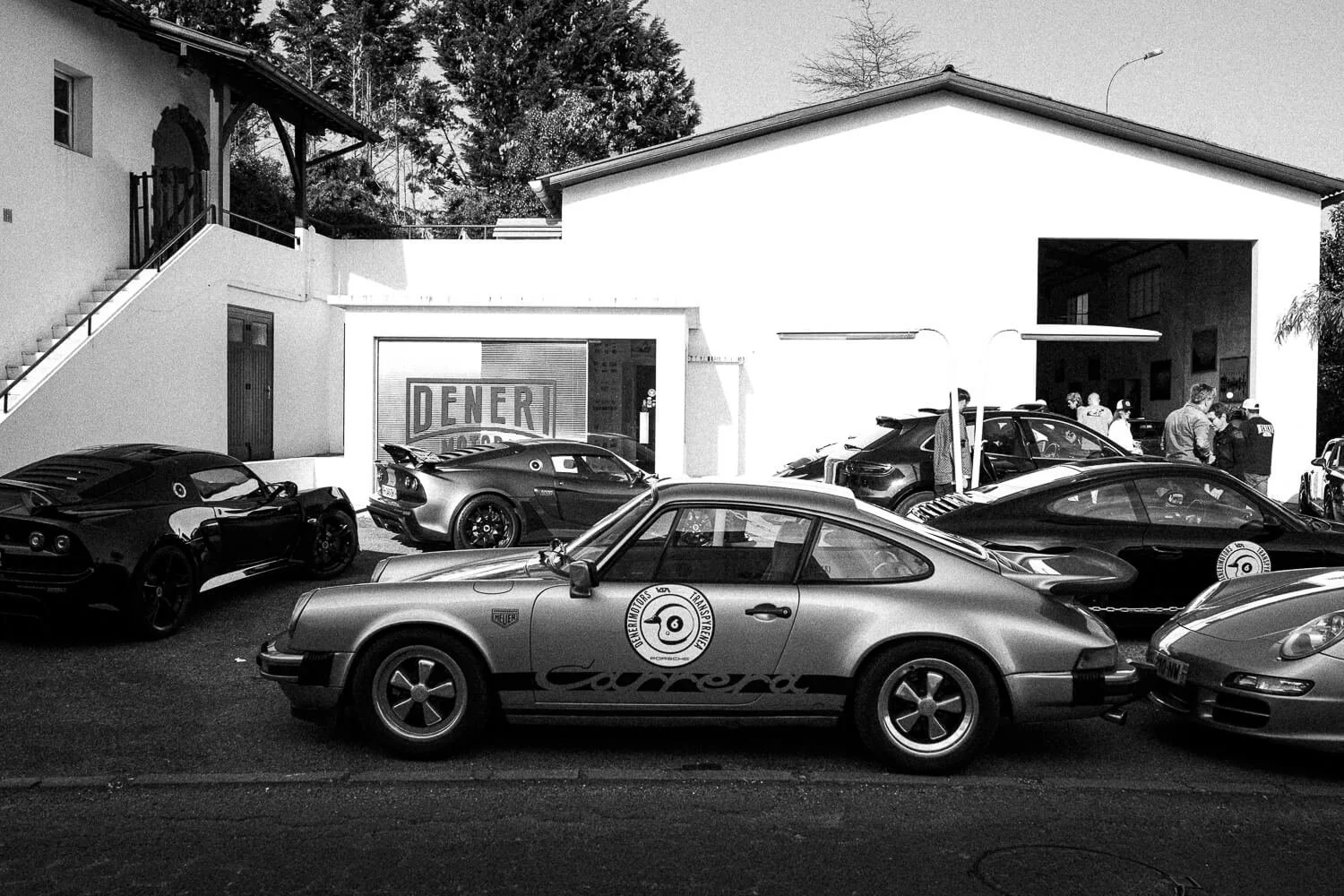 A parking lot with several cars, including a vintage Porsche, at a car exhibition. In the background, there is a white building with people gathered near the entrance and a sign that says 'DENER'.