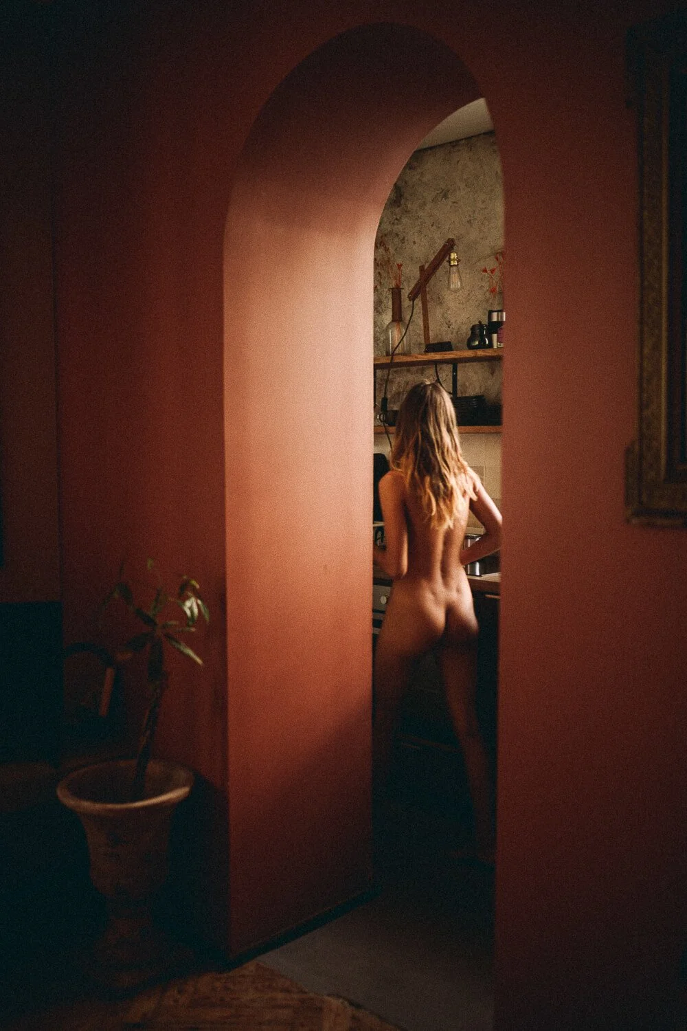 A woman with long hair stands nude in a kitchen, seen through an arched doorway, with warm lighting and rustic decor.