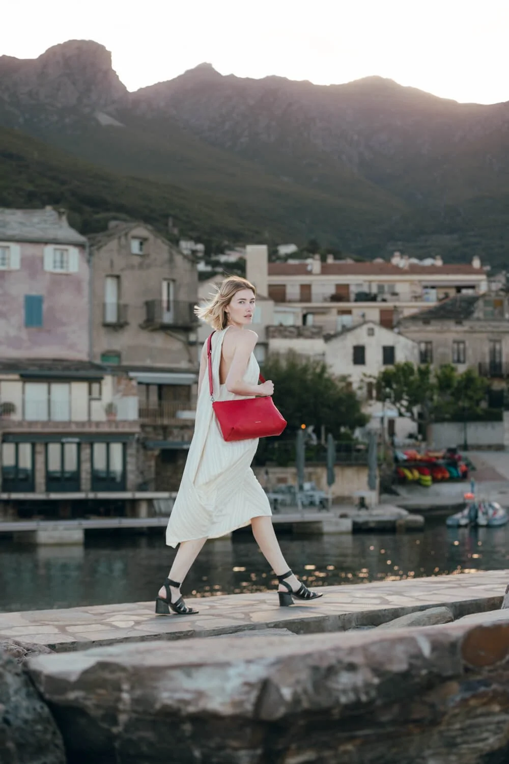 Woman in a white dress with black heels walking along a stone dock near water, carrying a red bag, with a small town and mountains in the background