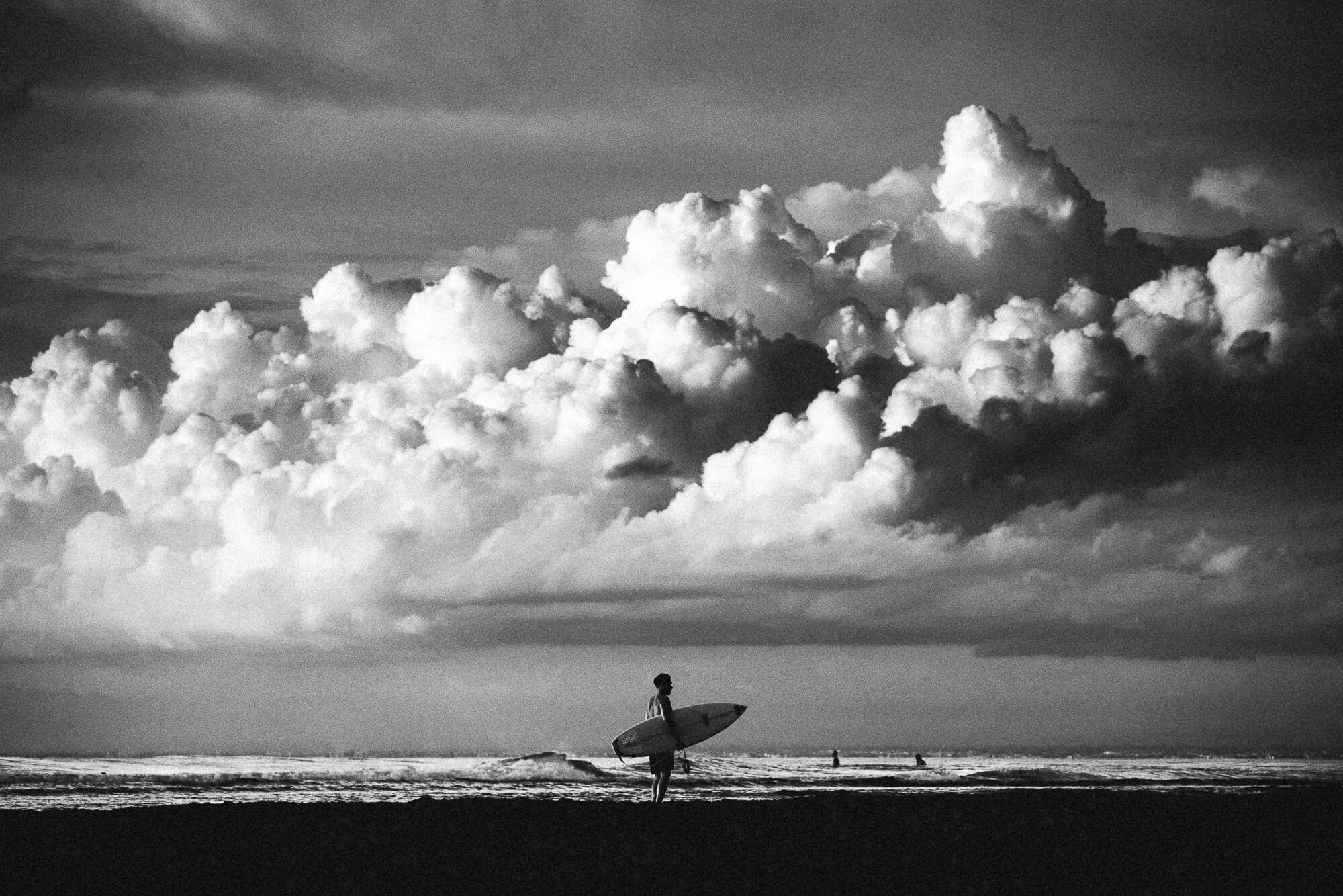 Un jeune sur la plage tenant une planche de surf, avec un ciel nuageux en noir et blanc