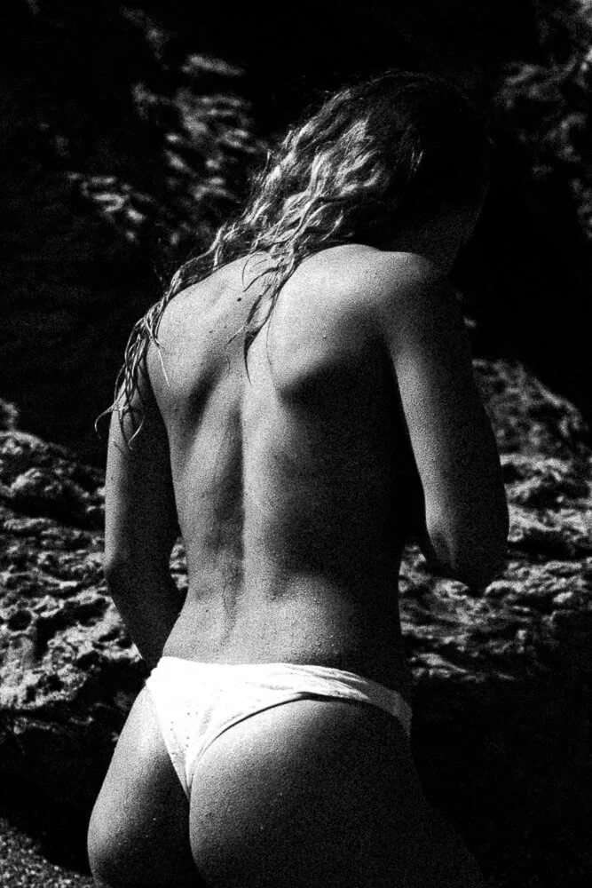 A black and white photo of a woman with long, wavy hair seen from behind, wearing only underwear, standing against a rocky background.