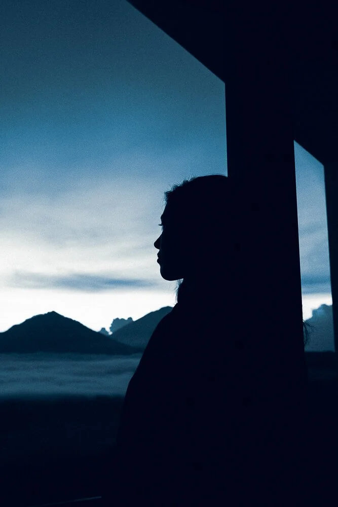 Silhouette of a woman standing by a window with mountains and cloudy sky in the background at dusk or dawn.