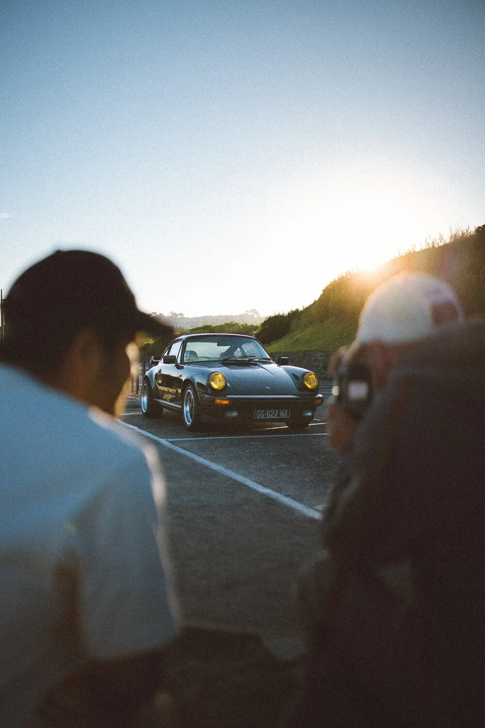 Two people sitting on the ground near a parked black classic Porsche car at sunset, with a landscape and hill in the background.