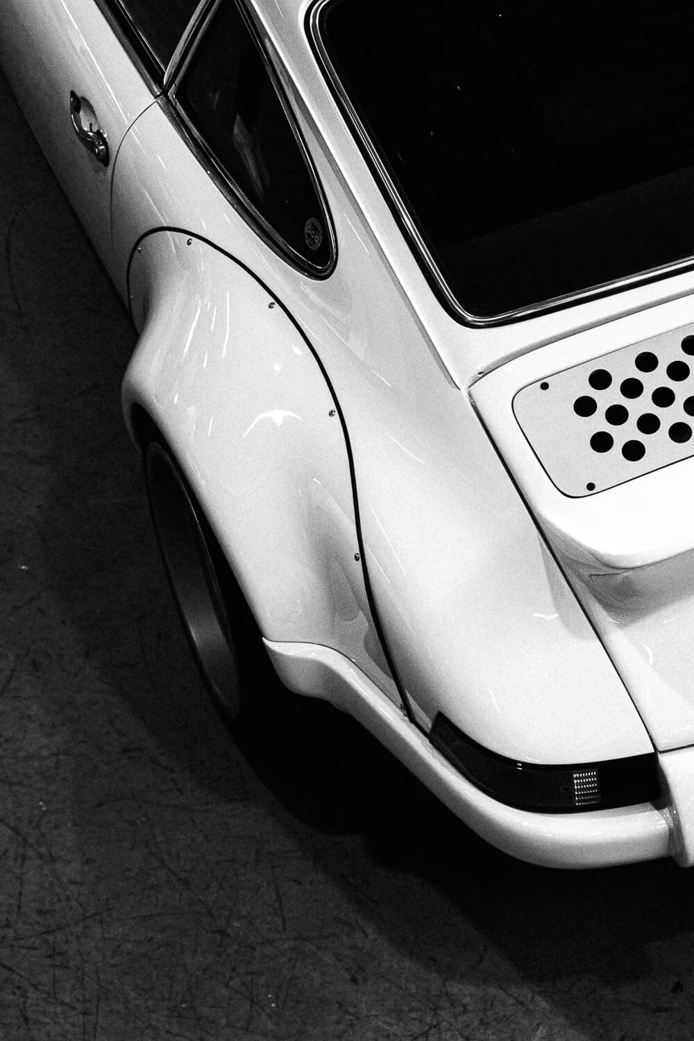 Close-up of a vintage white car with a damaged rear fender and black accents on the tail light and window, taken from a high angle in black and white.