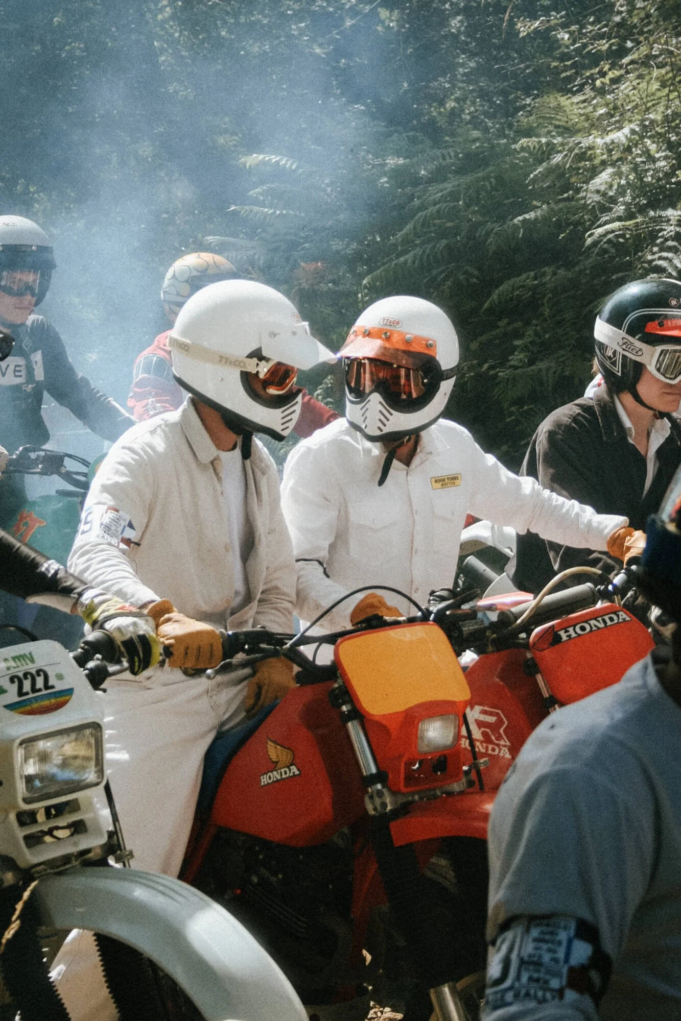 A group of motorcyclists wearing helmets and protective gear riding through a forested area, with some smoke in the background.