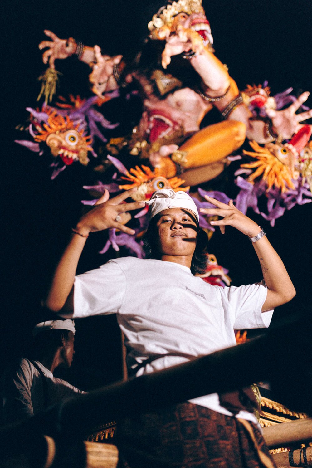 Une femme faisant un geste de paix avec les doigts, portant une robe blanche et un turban blanc, avec en arrière-plan une figure mythologique sur un décor coloré.