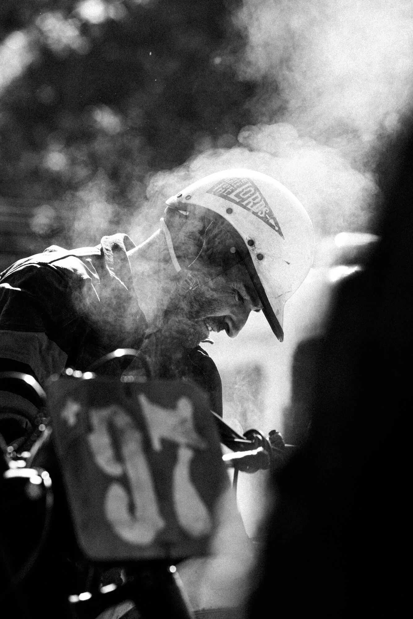 A motorcyclist wearing a helmet and protective gear, with the helmet bearing a logo, is riding through a cloud of dust or smoke. The photo is in black and white.