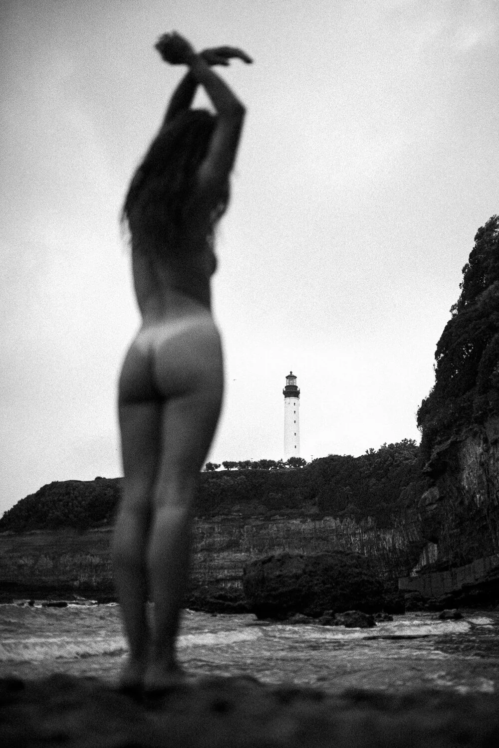 A woman standing on a beach, facing away, with a lighthouse in the distance in a black and white photograph.