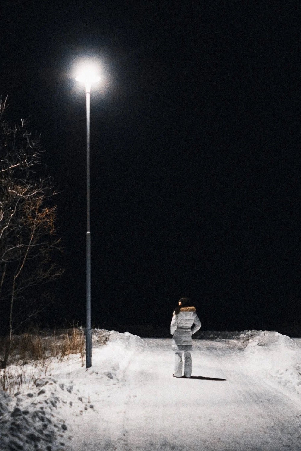 Une femme debout sur un chemin enneigé la nuit, éclairée par un lampadaire, avec un ciel noir et des arbres gelés à gauche.