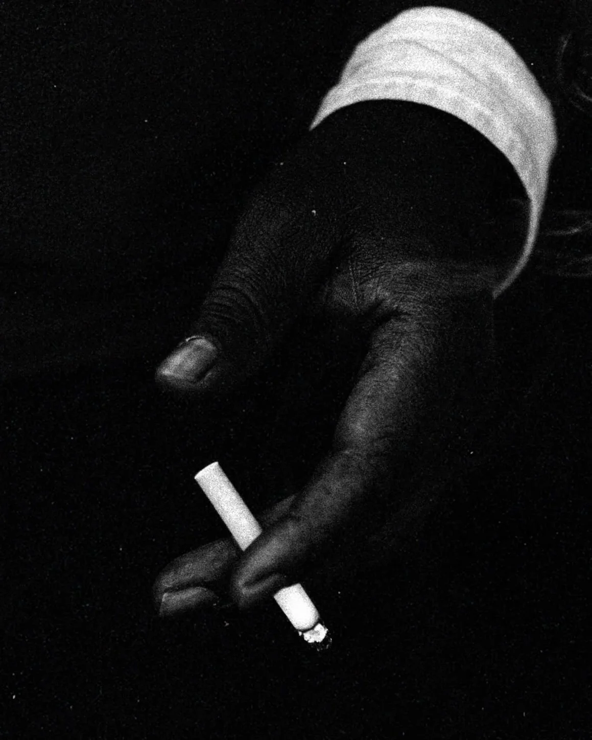 Une main noire tenant une cigarette dans un environnement sombre, avec une partie d'un vêtement blanc visible au poignet.