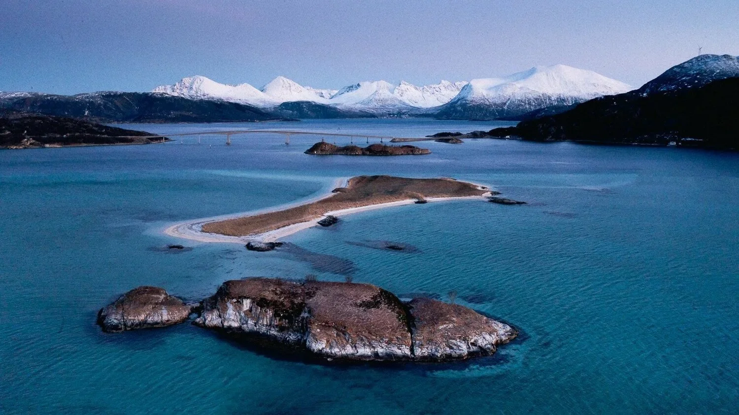Paysage de montagne enneigée, eau bleue, et petites îles ou rochers dans l'eau en Norvège.