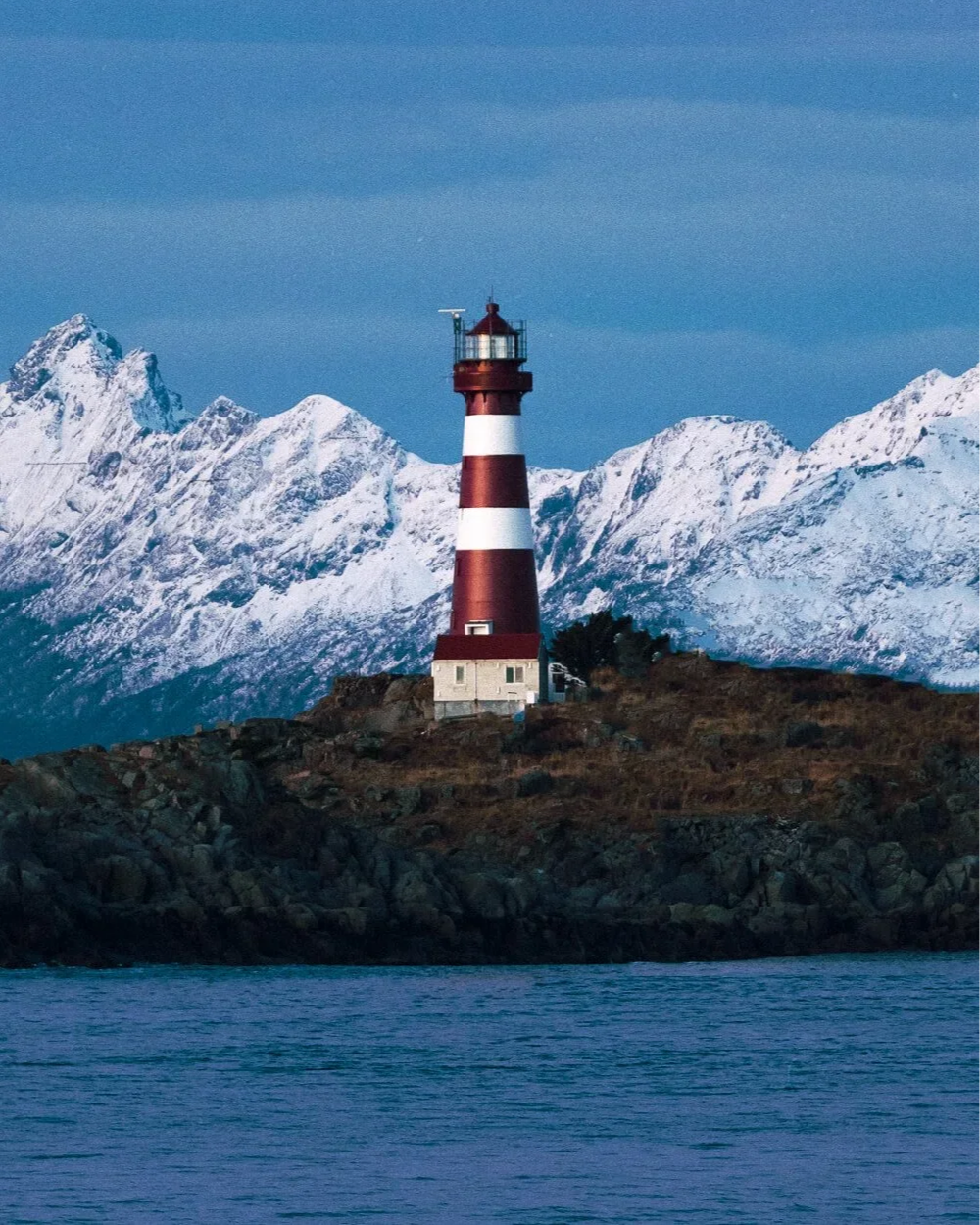 Phare rouge et blanc sur une île rocheuse, avec des montagnes enneigées en arrière-plan et l'eau au premier plan.