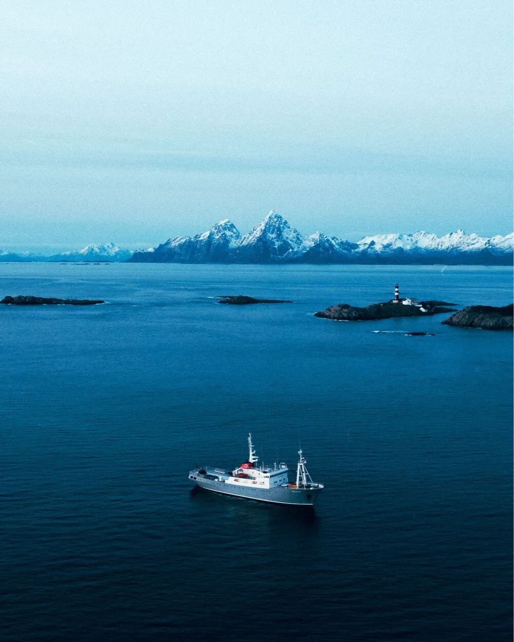 Un bateau navigue dans des eaux calmes avec une chaîne de montagnes enneigées à l'arrière-plan et un phare blanc et noir sur une petite île.