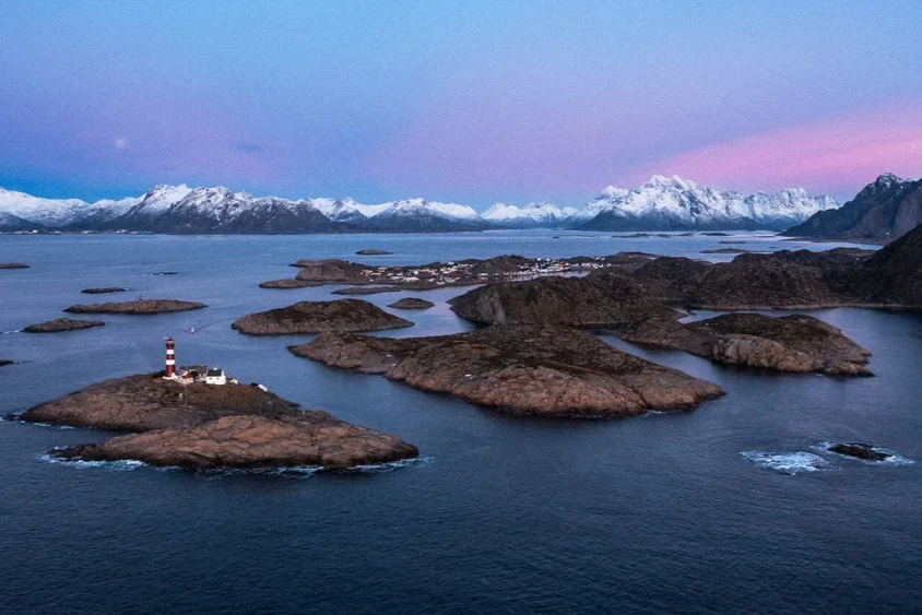 Paysage marin avec des îles rocheuses, un phare, montagnes enneigées en arrière-plan et un ciel coloré au crépuscule.
