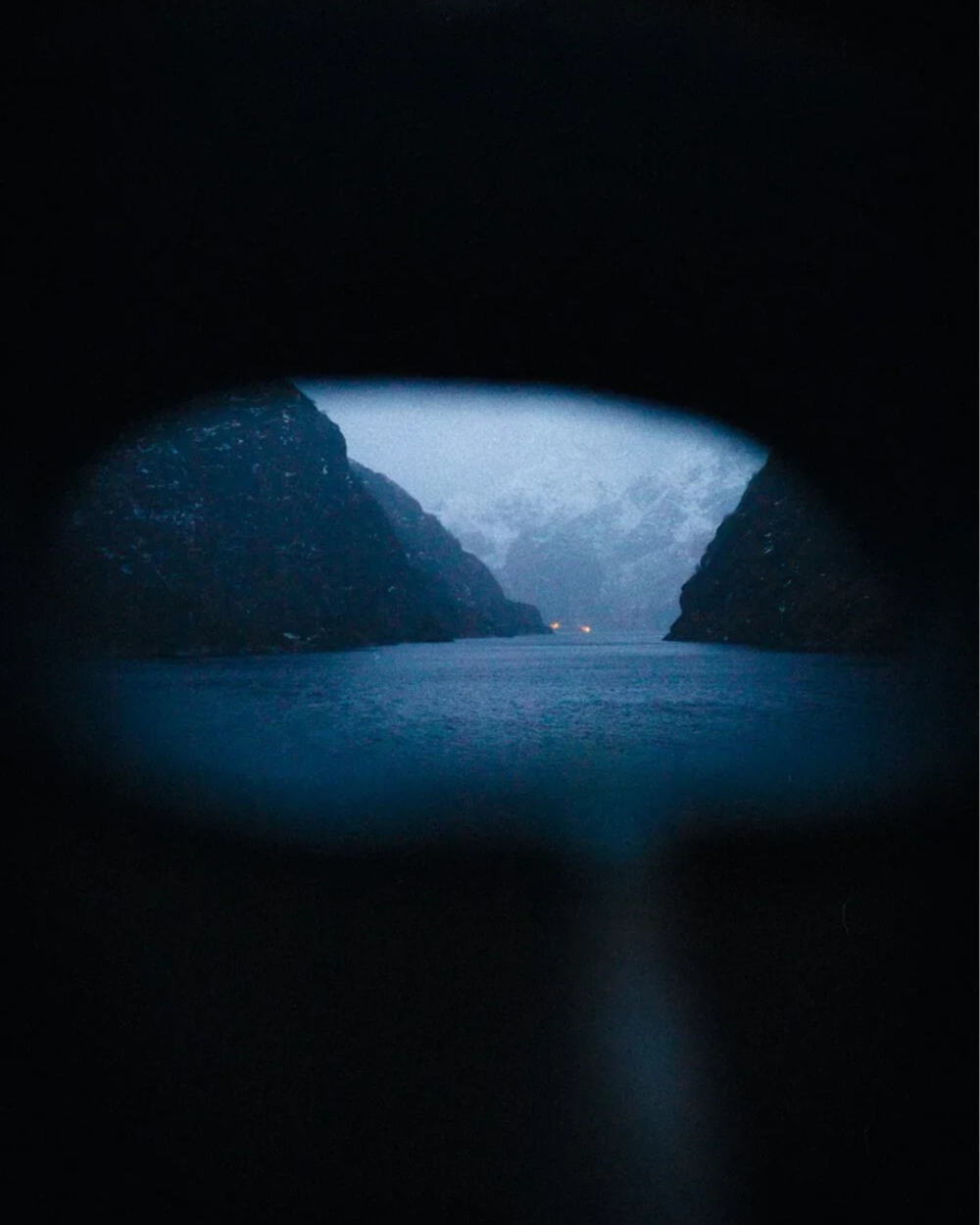 Vue d'une fjord à travers un trou d'une roche, avec des montagnes enneigées en arrière-plan, ambiance sombre modérée.