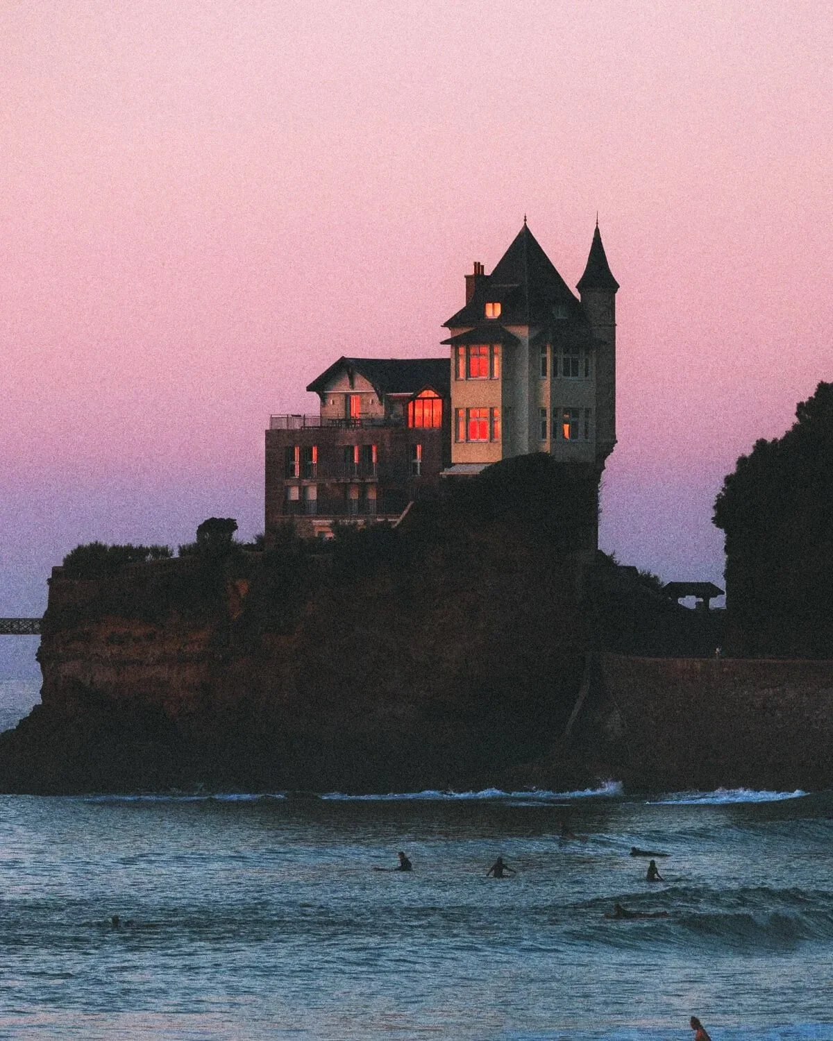 Château au bord de la mer au coucher du soleil avec des surfeurs sur l'eau.