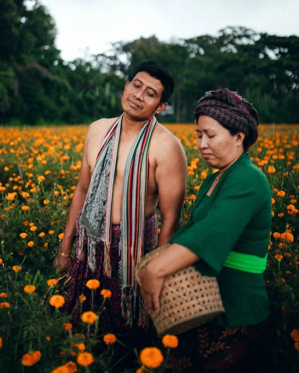 Deux personnes dans un champ de fleurs orange, une femme en vêtement traditionnel vert et un homme sans chemise portant une écharpe colorée.