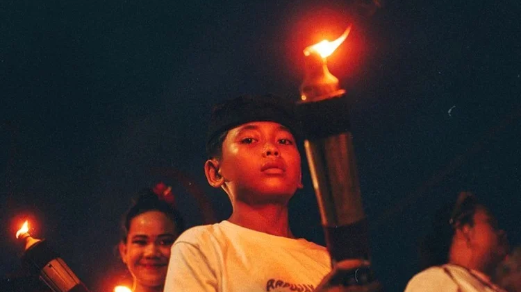Groupe d'enfants tenant des torches allumées lors d'une célébration nocturne, avec un enfant au centre regardant directement la caméra, arrière-plan sombre.
