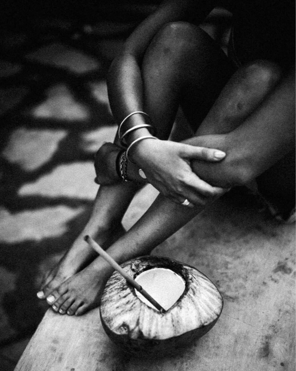 Photo en noir et blanc d'une personne assise, tenant ses jambes croisées d'une main, avec un bol de noix de coco à côté contenant une paille et un liquide. La personne porte des bracelets et est assise sur une surface en bois.