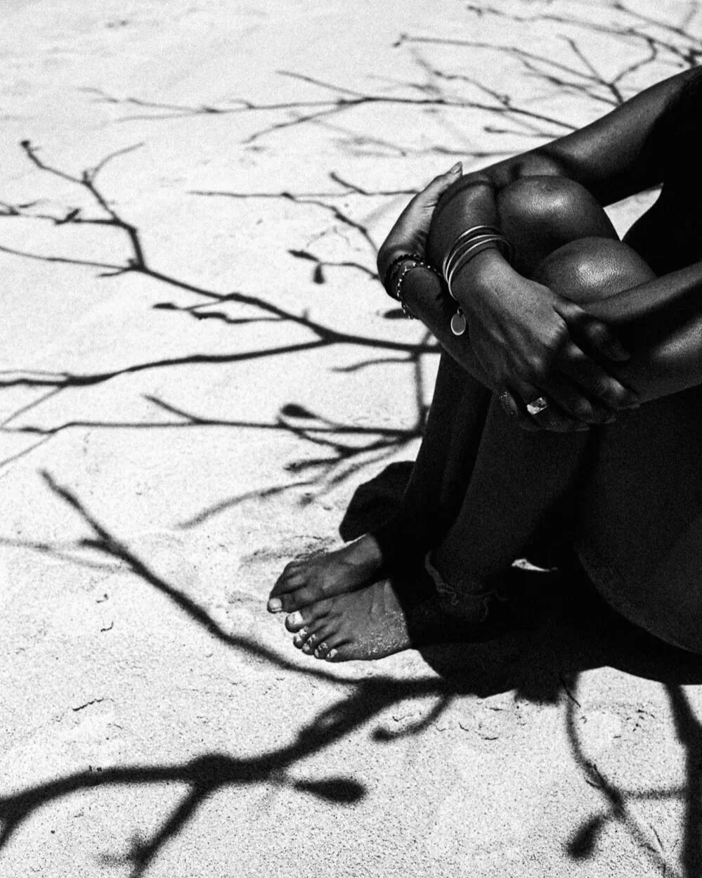 Une personne pieds nus assise sur du sable, entourée d'ombres de branches d'un arbre, en noir et blanc.