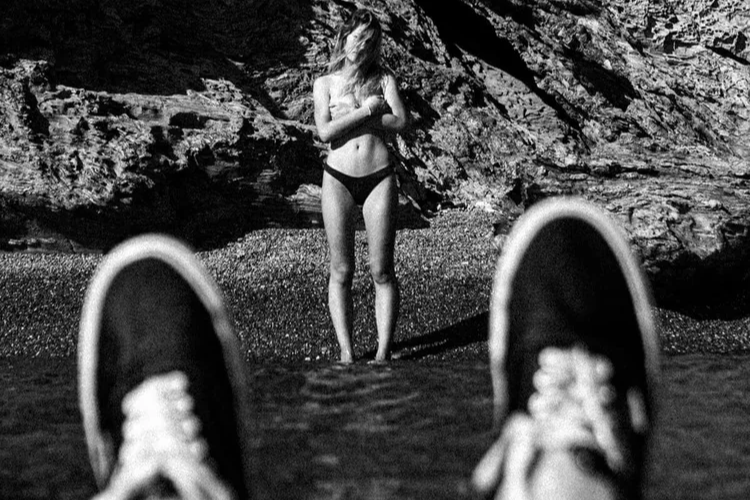 Photo en noir et blanc d'une femme debout sur une plage rocheuse, vue depuis une position allongée avec des chaussures visibles au premier plan.