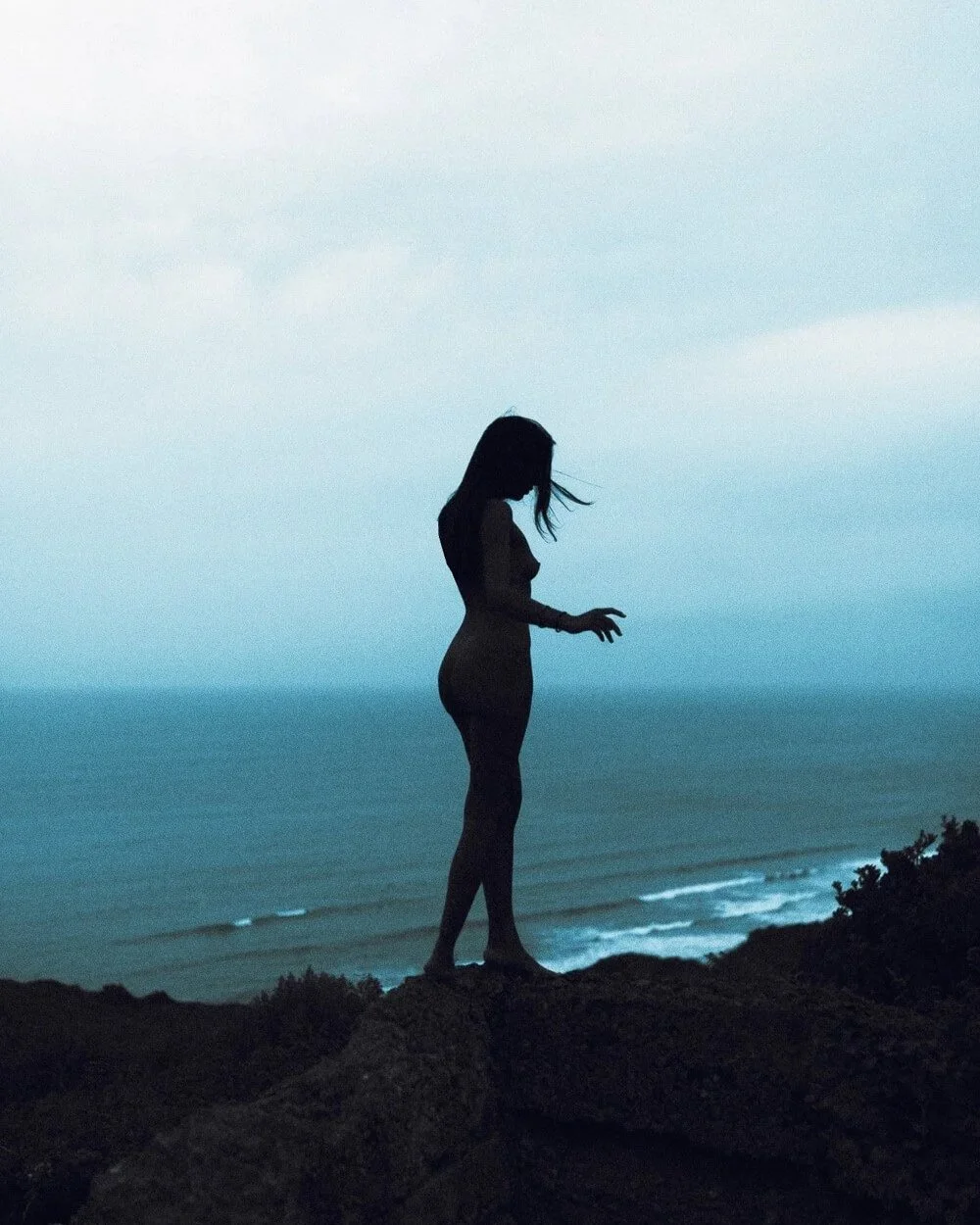 Silhouette of a woman standing on rocks overlooking the ocean during twilight, with a cloudy sky above.