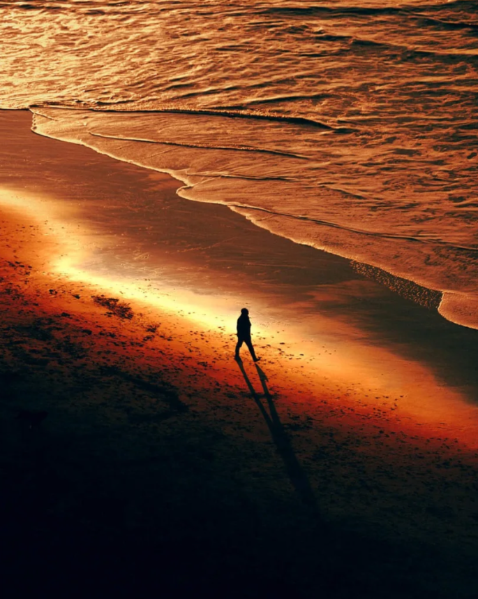 Person walking alone on a beach at sunset with long shadows and gentle waves.