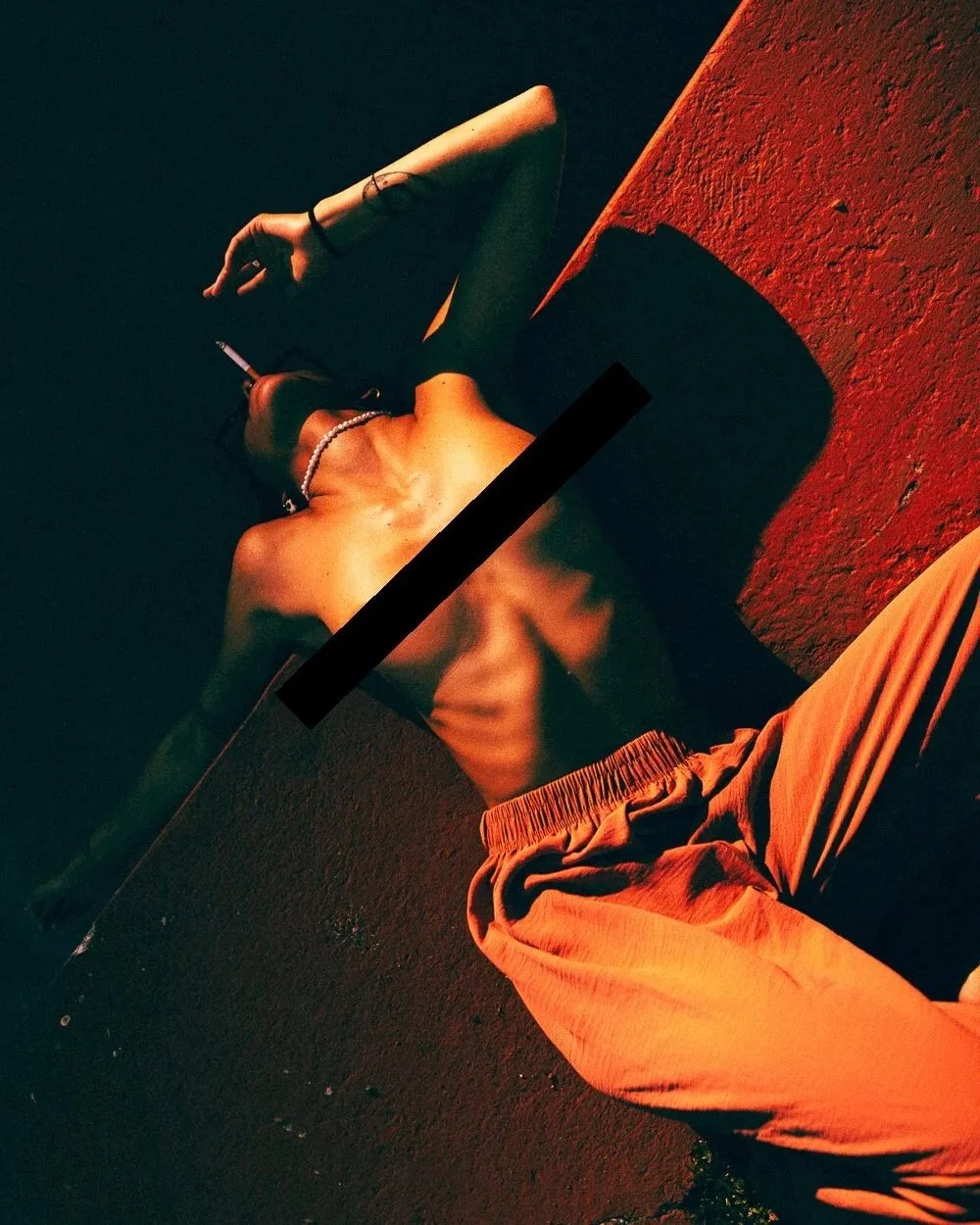 Person lying on the ground under red lighting, wearing orange shorts, holding a cigarette, and wearing a pearl necklace and bracelet.