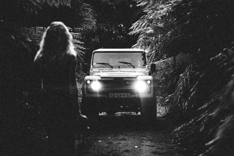 Une femme marche sur un chemin de forêt sombre, avec un véhicule dont les phares illuminent la route, entouré d'arbres et de feuillages.