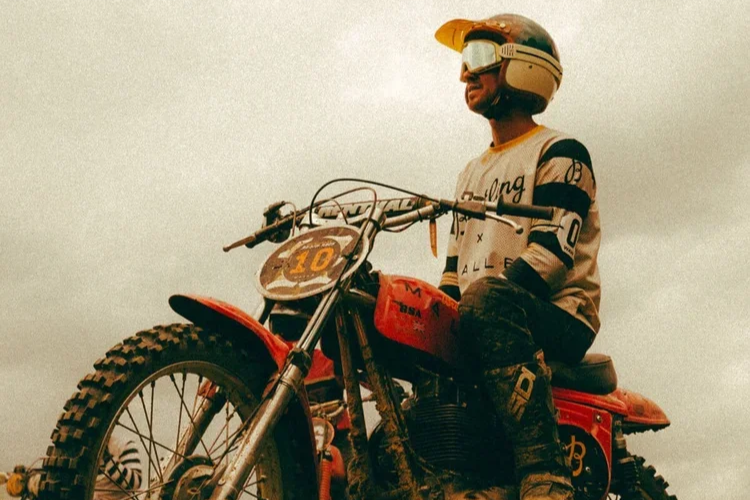 A person wearing a helmet and goggles sitting on a red motorcycle outdoors under an overcast sky.