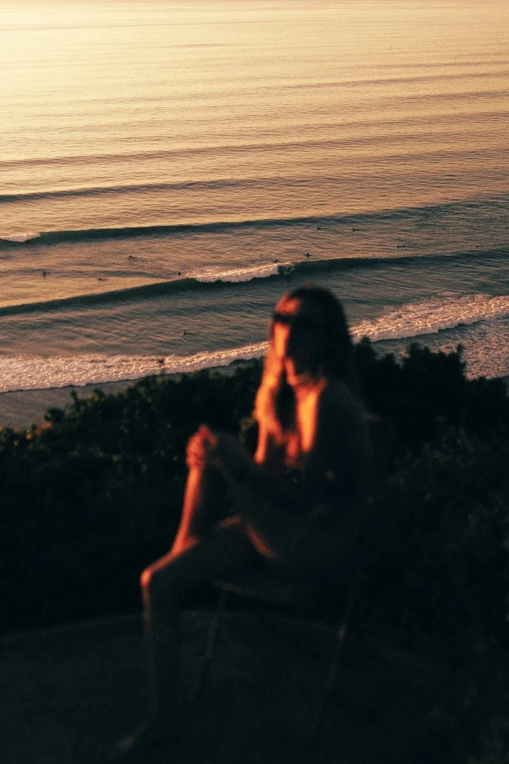 A person sitting outdoors during sunset, overlooking the ocean with visible waves and surfers in the water.