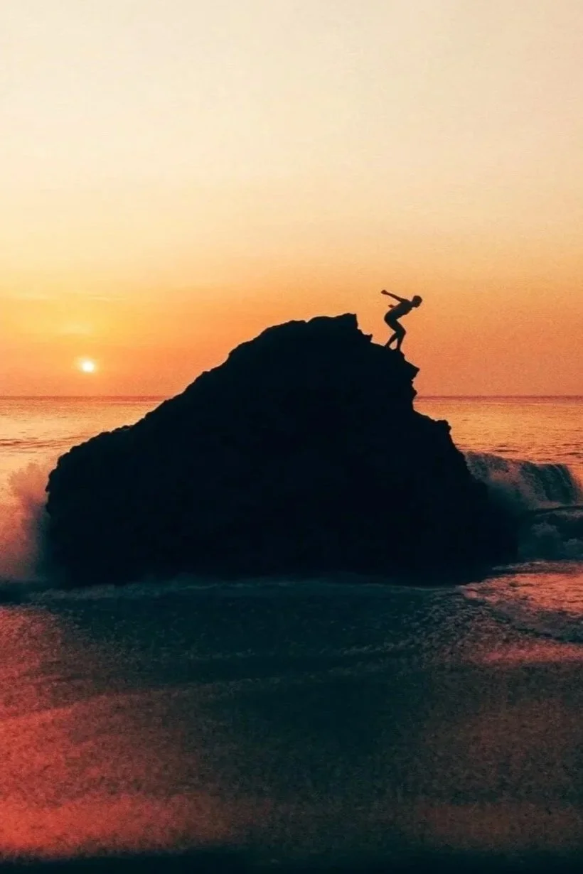Person climbing on a large rock at the beach during sunset.