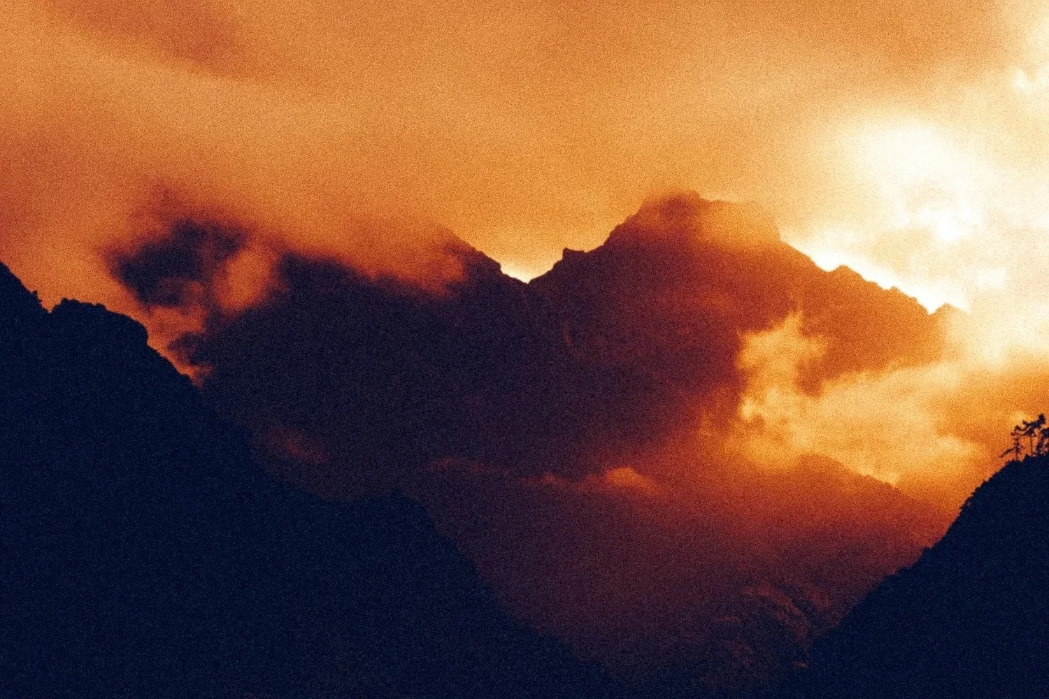 Mountains with dark peaks beneath an orange sky, filled with thick clouds and smoke.