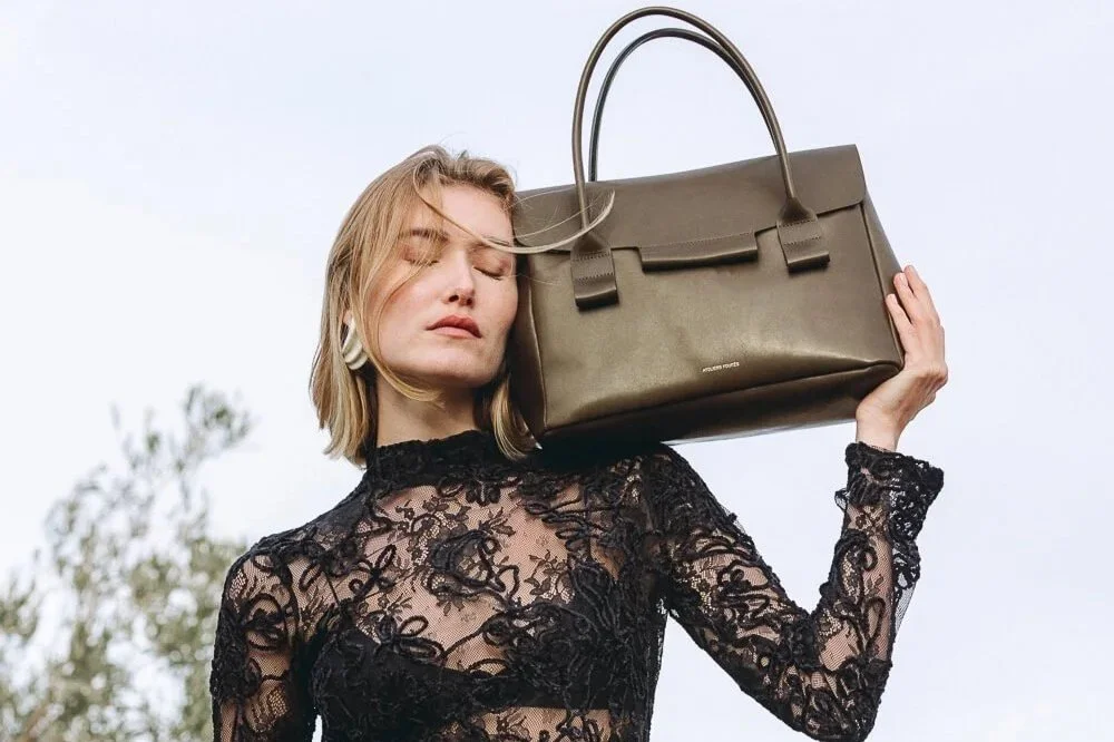 Woman in a black lace top carrying a large olive green handbag on her shoulder against a light sky background.