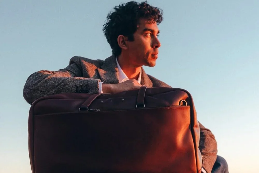 A young man in a gray blazer sitting outdoors with a large black suitcase, looking into the distance at sunset or sunrise.