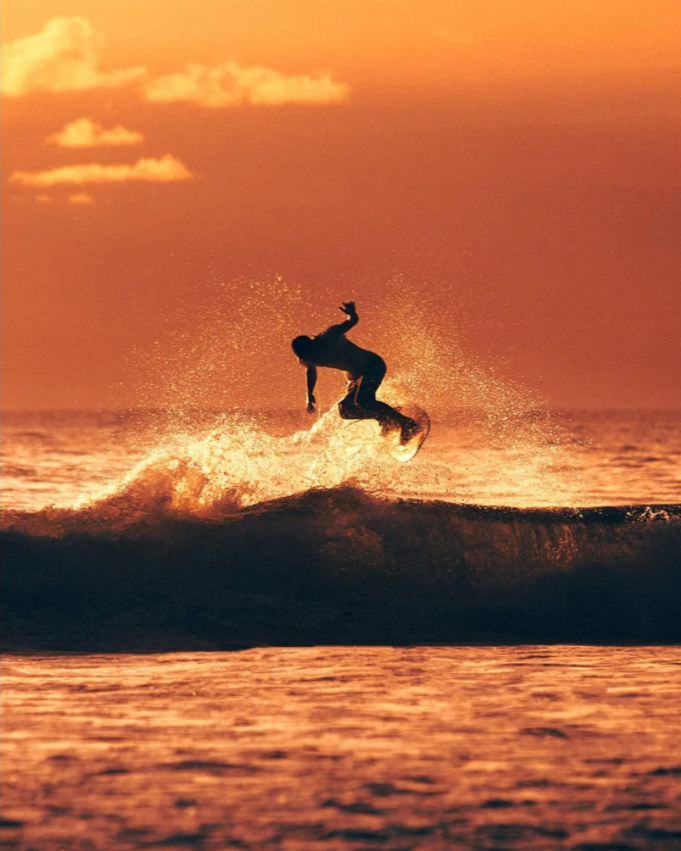 A person surfing on a wave during sunset with an orange sky and some clouds.
