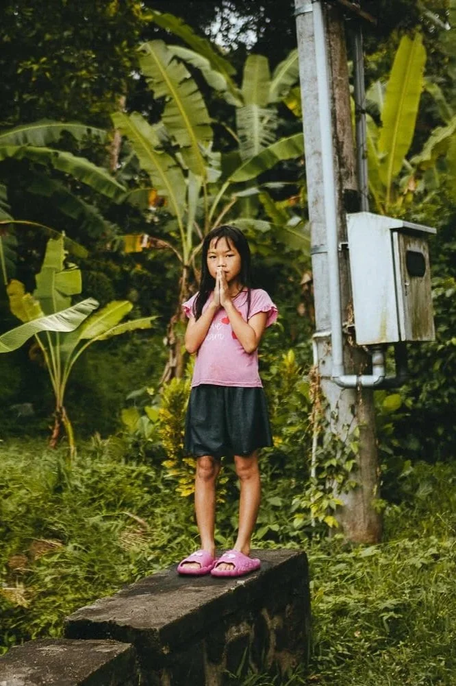 Young girl standing on a stone ledge with palms pressed together in prayer, surrounded by lush green tropical plants and banana trees.