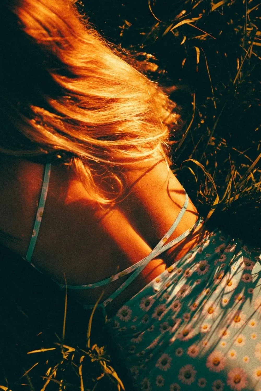 Close-up of a woman lying in grass, wearing a daisy-patterned dress, with sunlight illuminating her blonde hair.