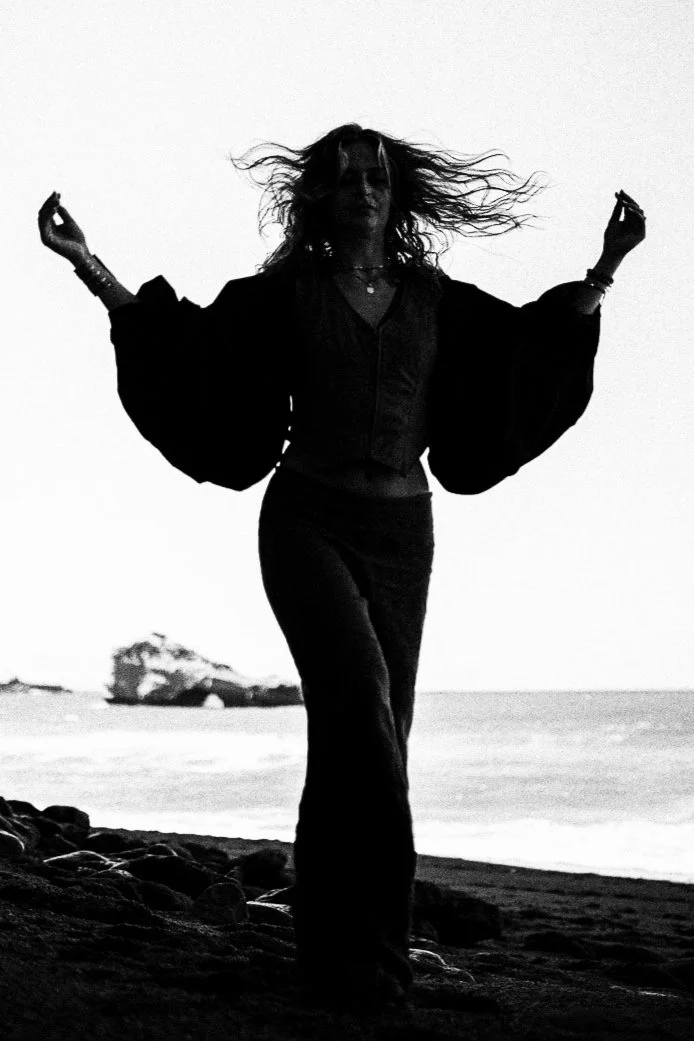 A person with curly hair dancing on a beach, silhouetted against the sky, with rocks and an ocean in the background.