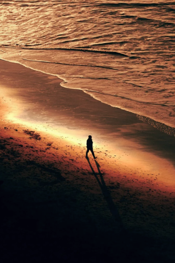 Person walking alone on a beach at sunset with long shadows and gentle waves.