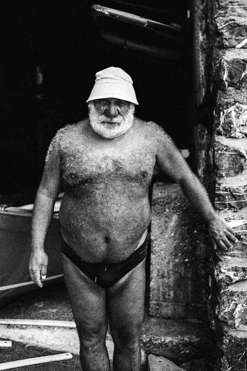 Homme âgé barbu, portant un chapeau de pêcheur, torse nu et en sous-vêtements, debout à côté d’un mur en pierre, dans un environnement intérieur.