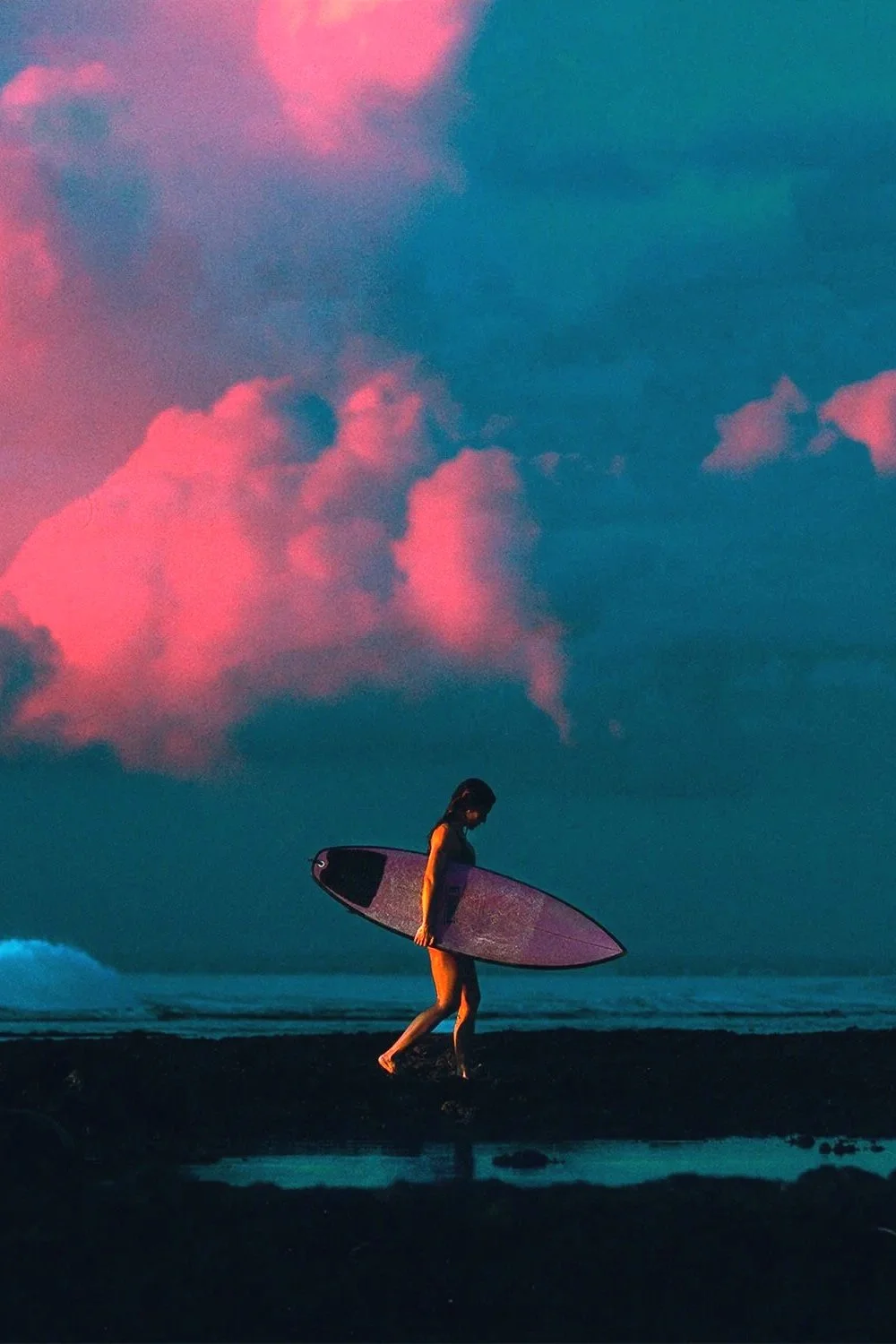 Une personne marchant sur la plage avec une planche de surf au coucher du soleil, ciel nuageux teinté de rose.