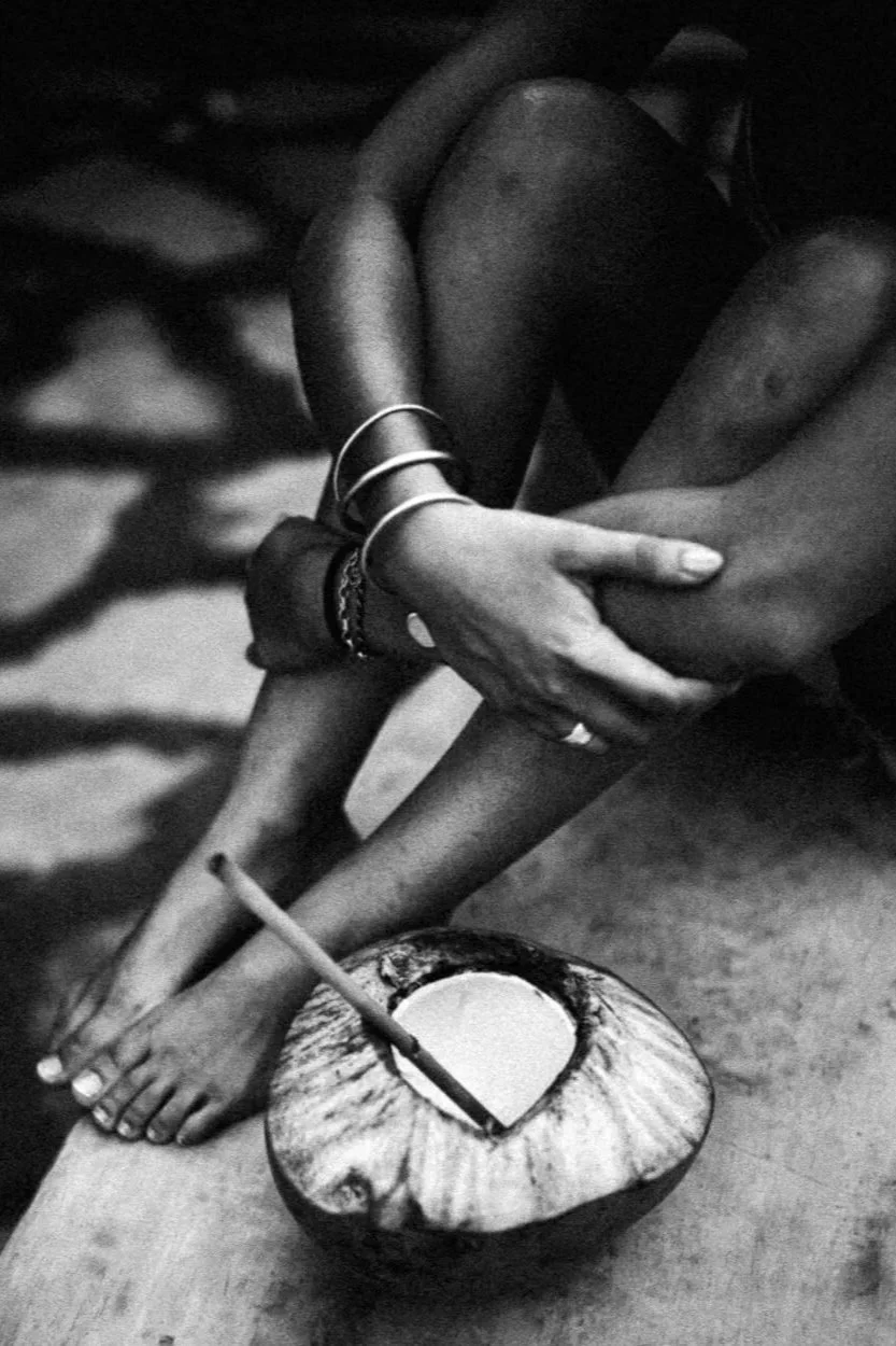 Photo en noir et blanc d'une personne assise, tenant ses jambes croisées d'une main, avec un bol de noix de coco à côté contenant une paille et un liquide. La personne porte des bracelets et est assise sur une surface en bois.