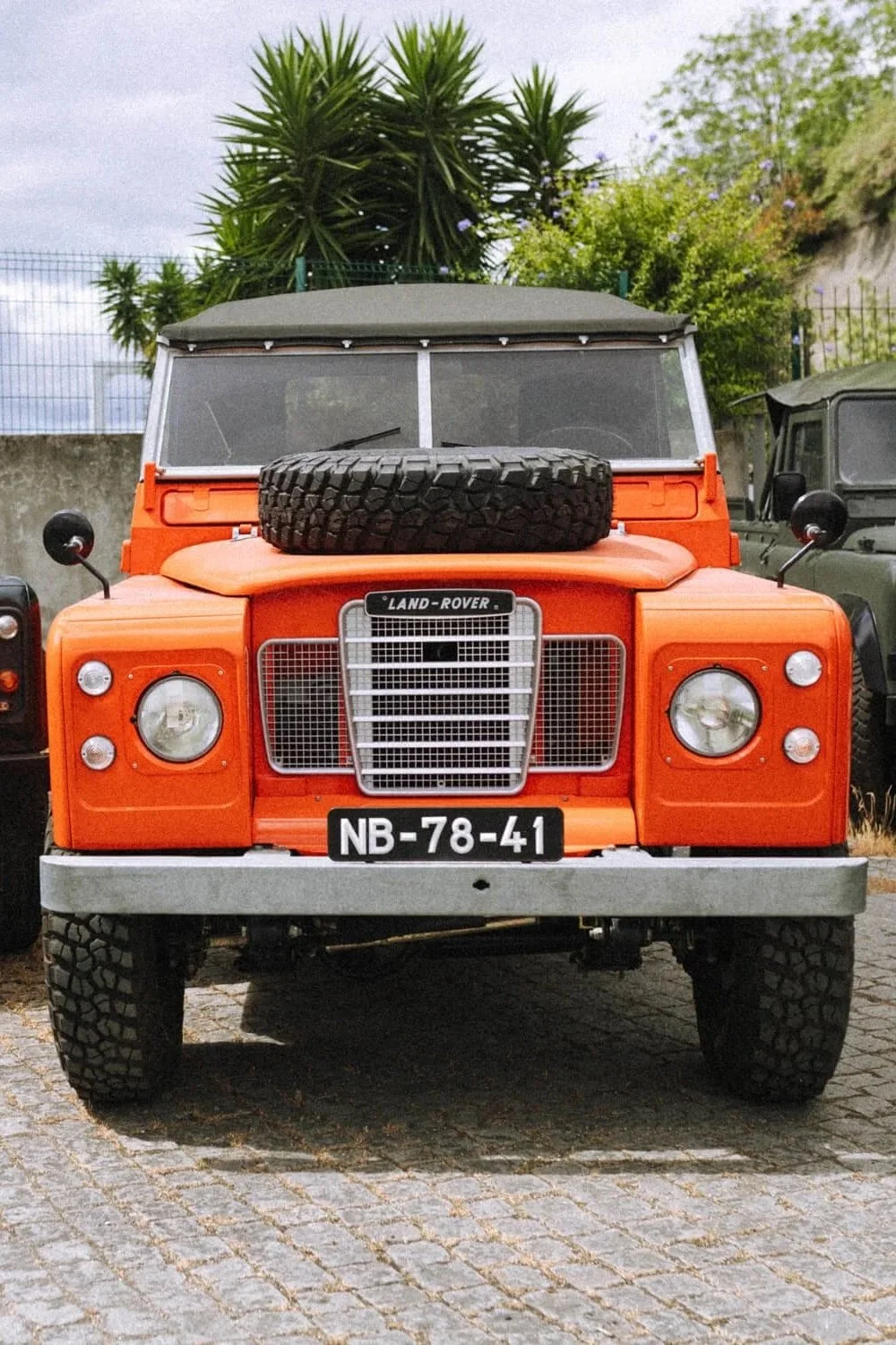 Un véhicule tout-terrain Land Rover orange avec un pneu de secours sur le capot, stationné sur une allée pavée, entouré de végétation et d'autres véhicules.