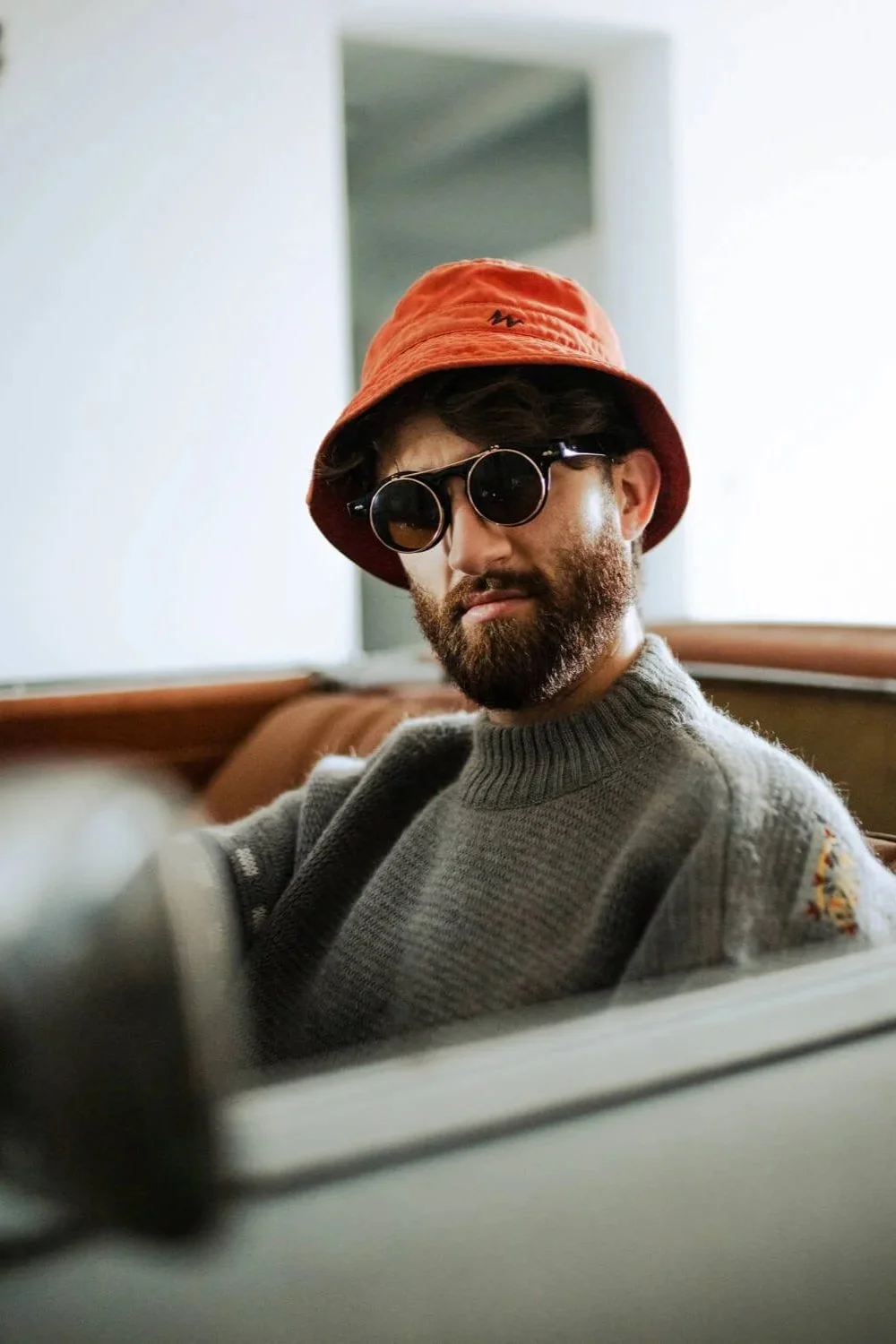 Un homme barbu portant un chapeau orange et des lunettes de soleil assis dans une voiture, vêtu d'un pull gris.