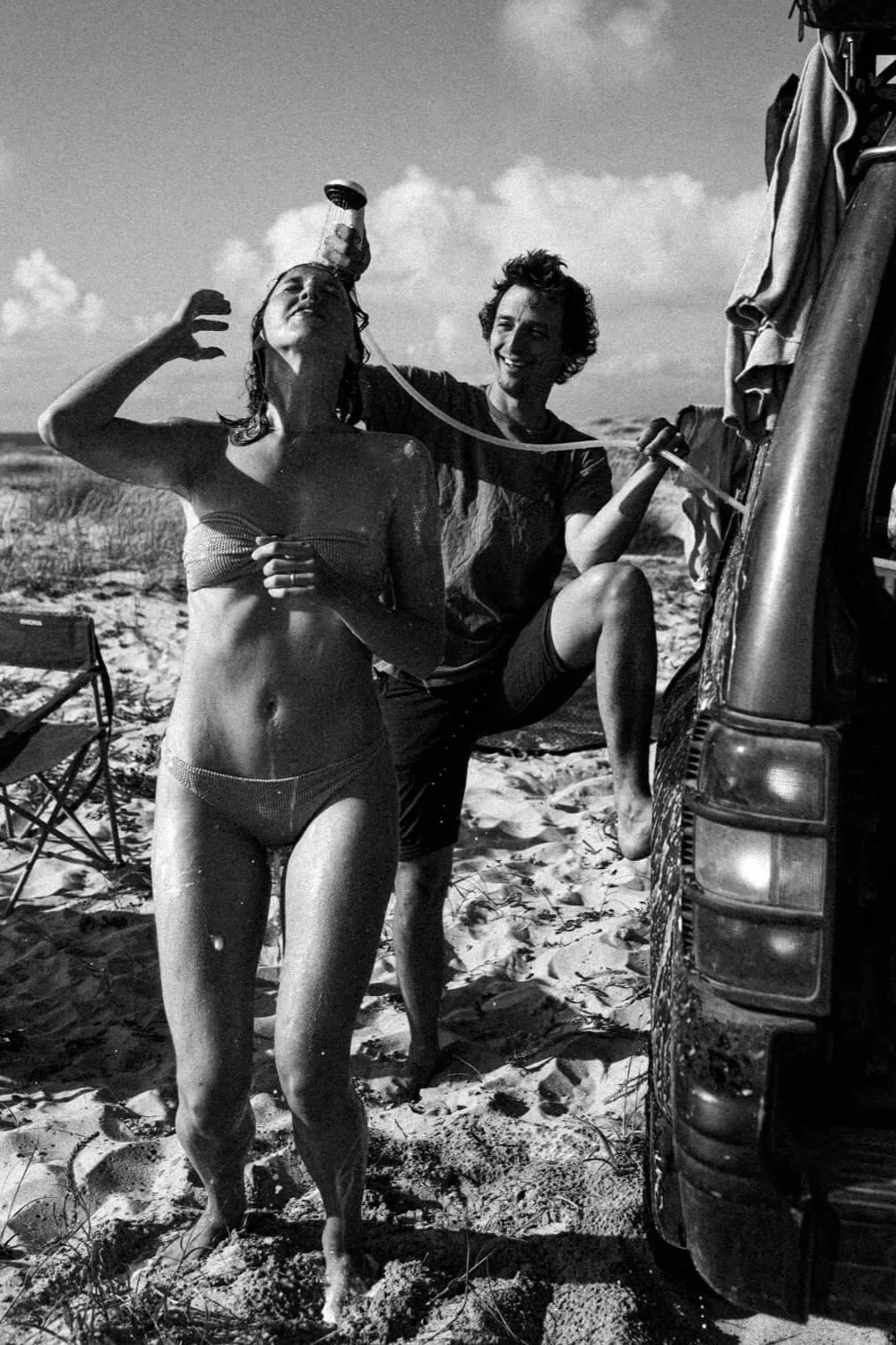 Deux personnes sur une plage. Une femme en maillot de bain se douche à l'extérieur, à l'aide d'une douche portable. Un homme tient la douche, debout près d'une voiture. Scène en noir et blanc.