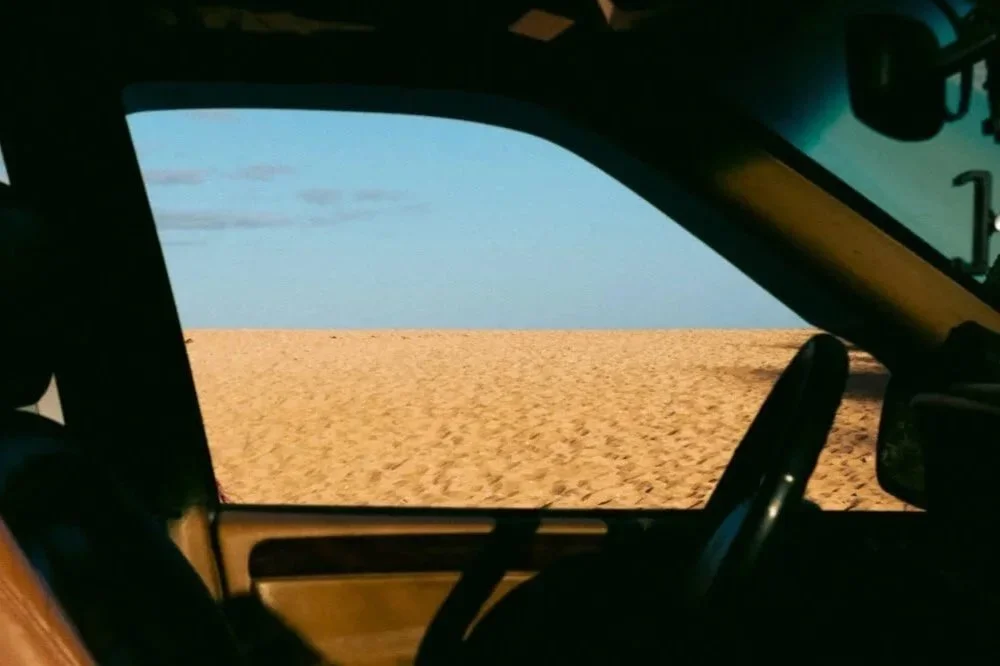Vue depuis l'intérieur d'une voiture vers un paysage désertique avec du sable et un ciel bleu.