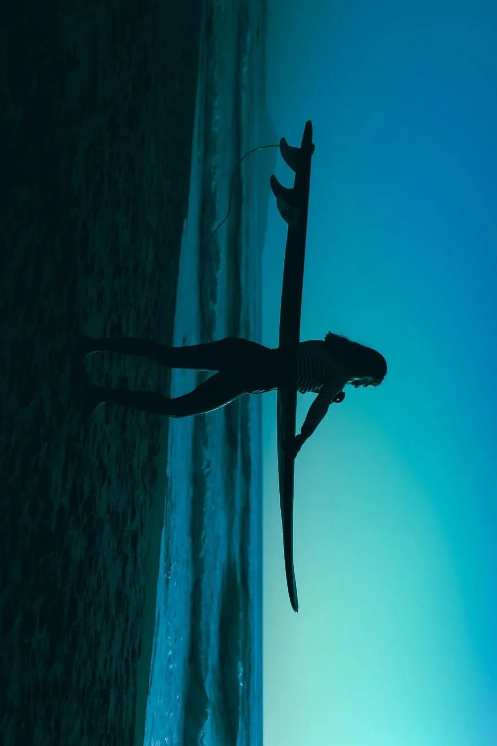Silhouette of a woman holding a paddleboard at the beach during sunset with the ocean in the background.
