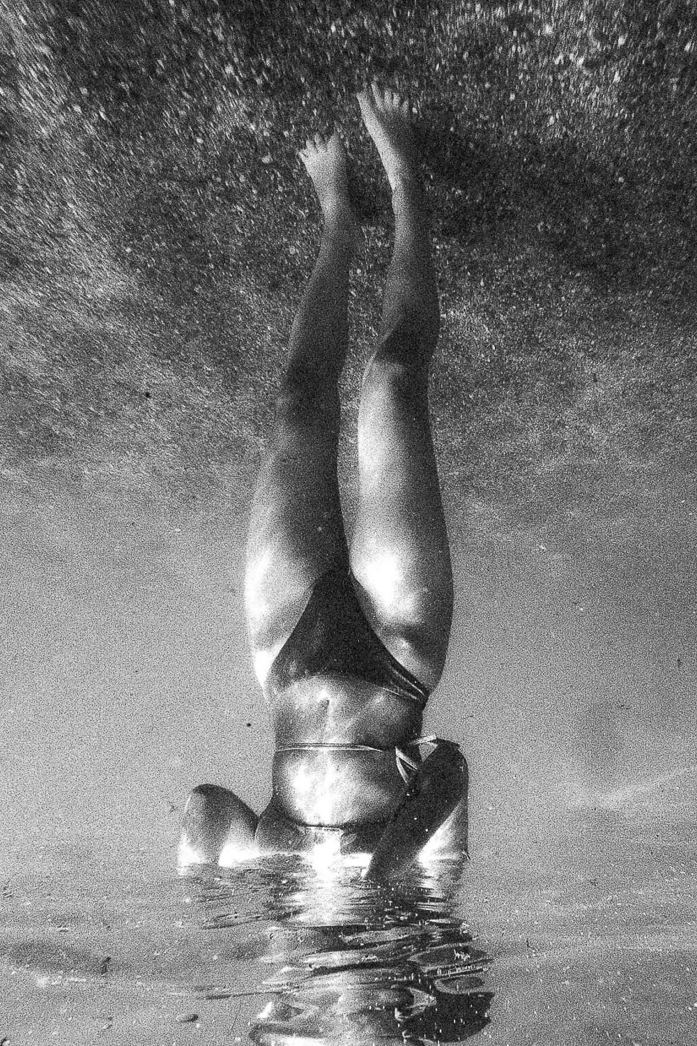 A metallic sculpture of a human figure lying face down, with legs extended and head resting on one arm, located on a beach with wet sand and a cloudy sky.